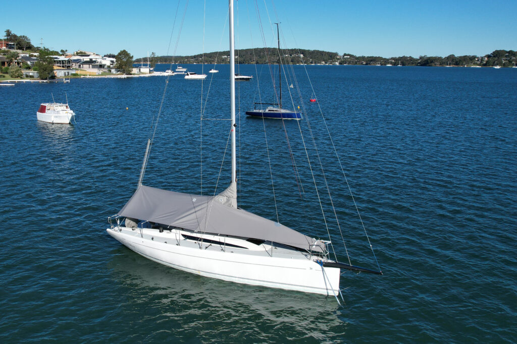 Custom overall protection cover — Italia Yachts 9.98 on mooring, Lake Macquarie Aerial drone view of an Italia Yachts 9.98 Bellissima on a swing mooring in Lake Macquarie fitted with a custom grey WeatherMax overall protection cover by Ocean Covers, with other moored boats and foreshore in the background
